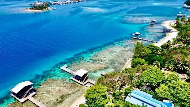 Vista aérea de una zona costera con aguas azul turquesa, una pequeña isla con construcciones y una playa con un muelle.