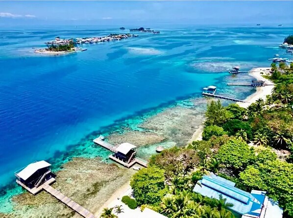 Une vue aérienne d’une zone côtière avec des eaux d’un bleu limpide, une petite île dotée de quelques constructions et une plage avec une jetée.