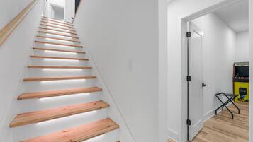 Una escalera con escalones de madera y paredes blancas que conduce a una habitación con una puerta y una máquina de juegos.