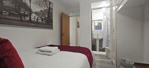 A hotel room with a bed, a red blanket, a white towel, a painting of a cityscape, and a bathroom visible through an open door. - Chiswick Residence (London)