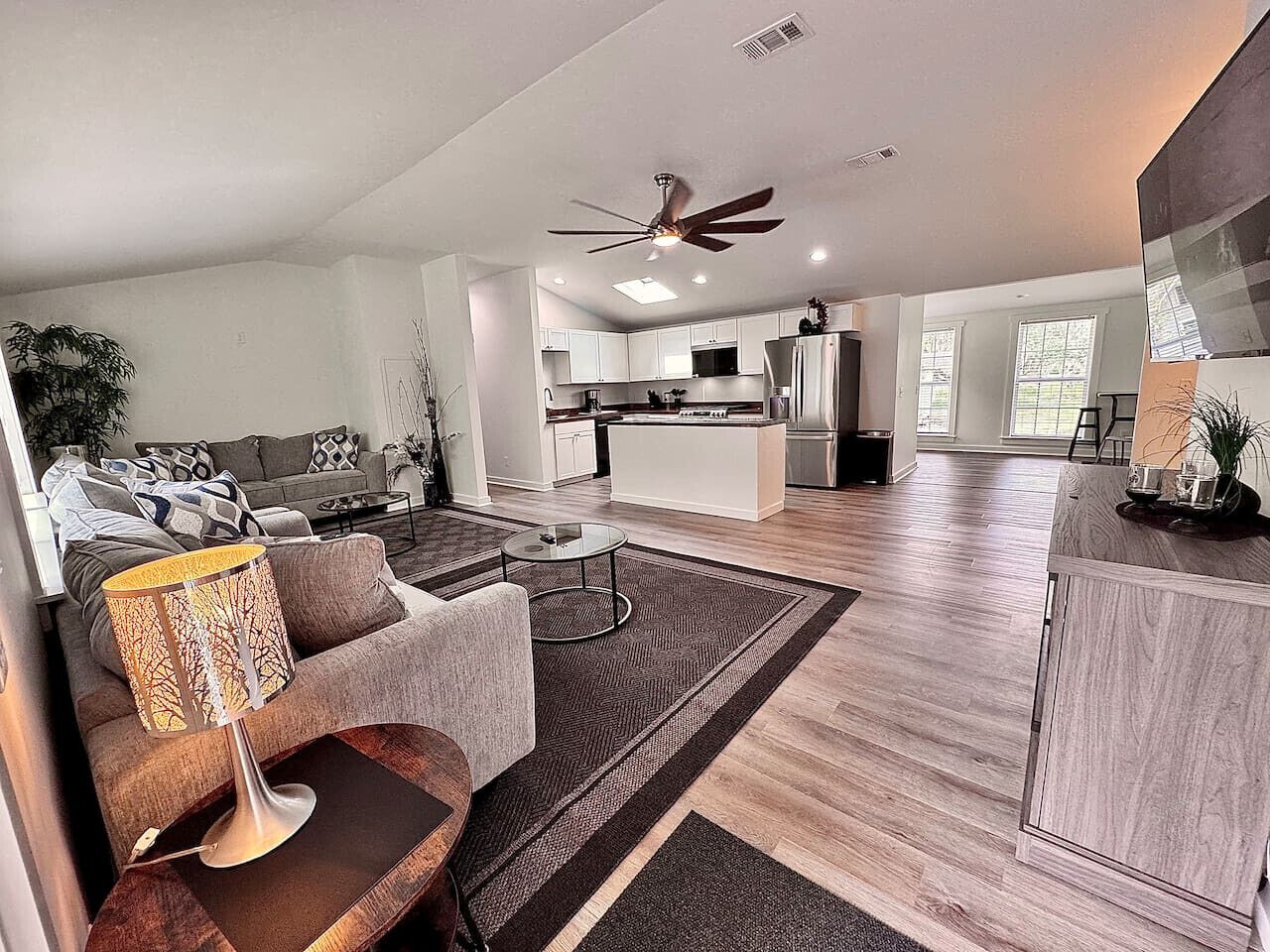 A modern living room with a sofa, coffee table, and lamp, leading to a kitchen with white cabinets and a dining area.