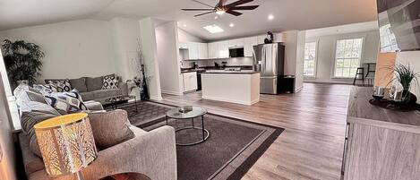 A modern living room with a sofa, coffee table, and lamp, leading to a kitchen with white cabinets and a dining area.