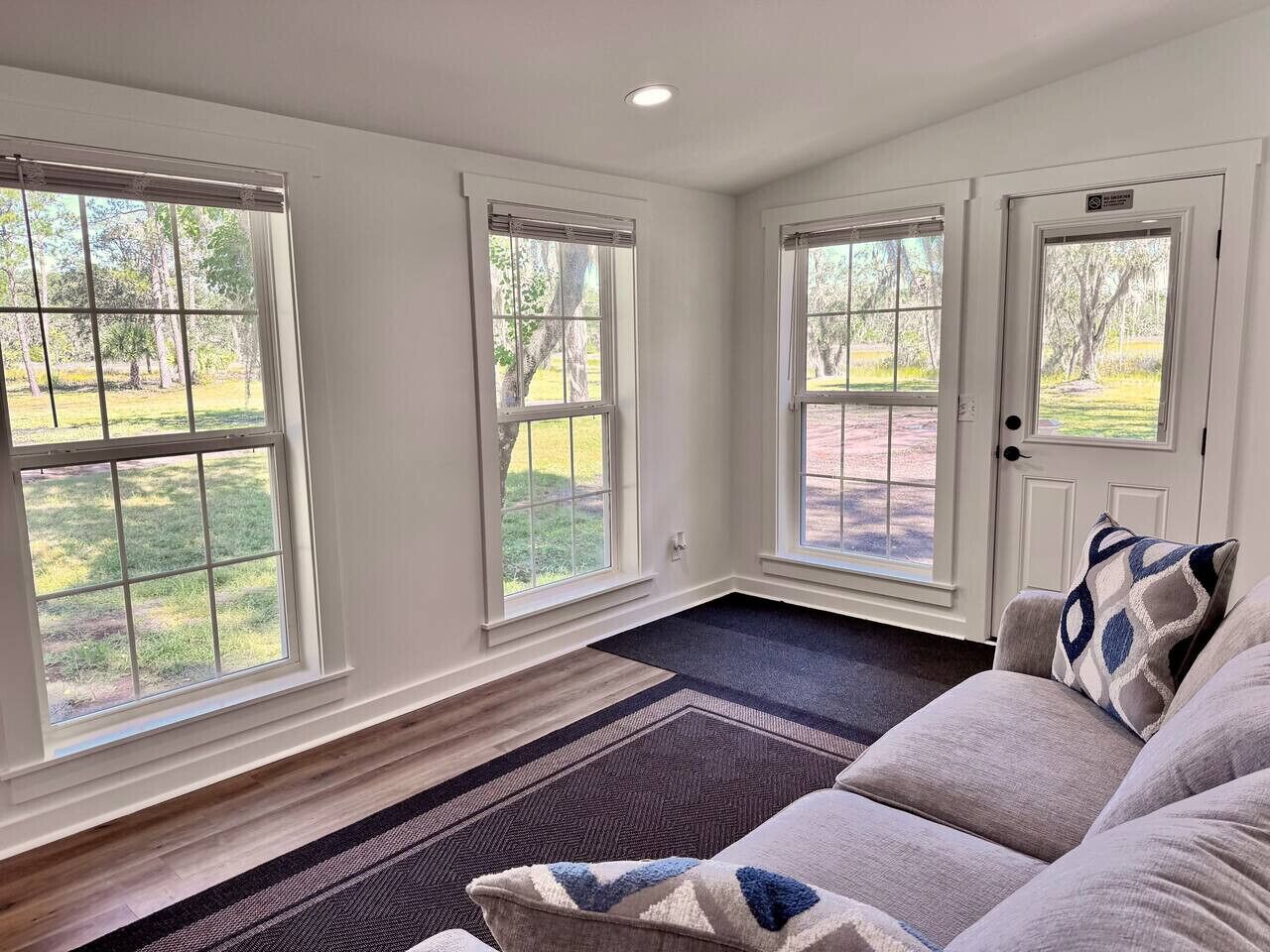 A room with large windows, a door, a patterned rug, and a sofa with pillows.