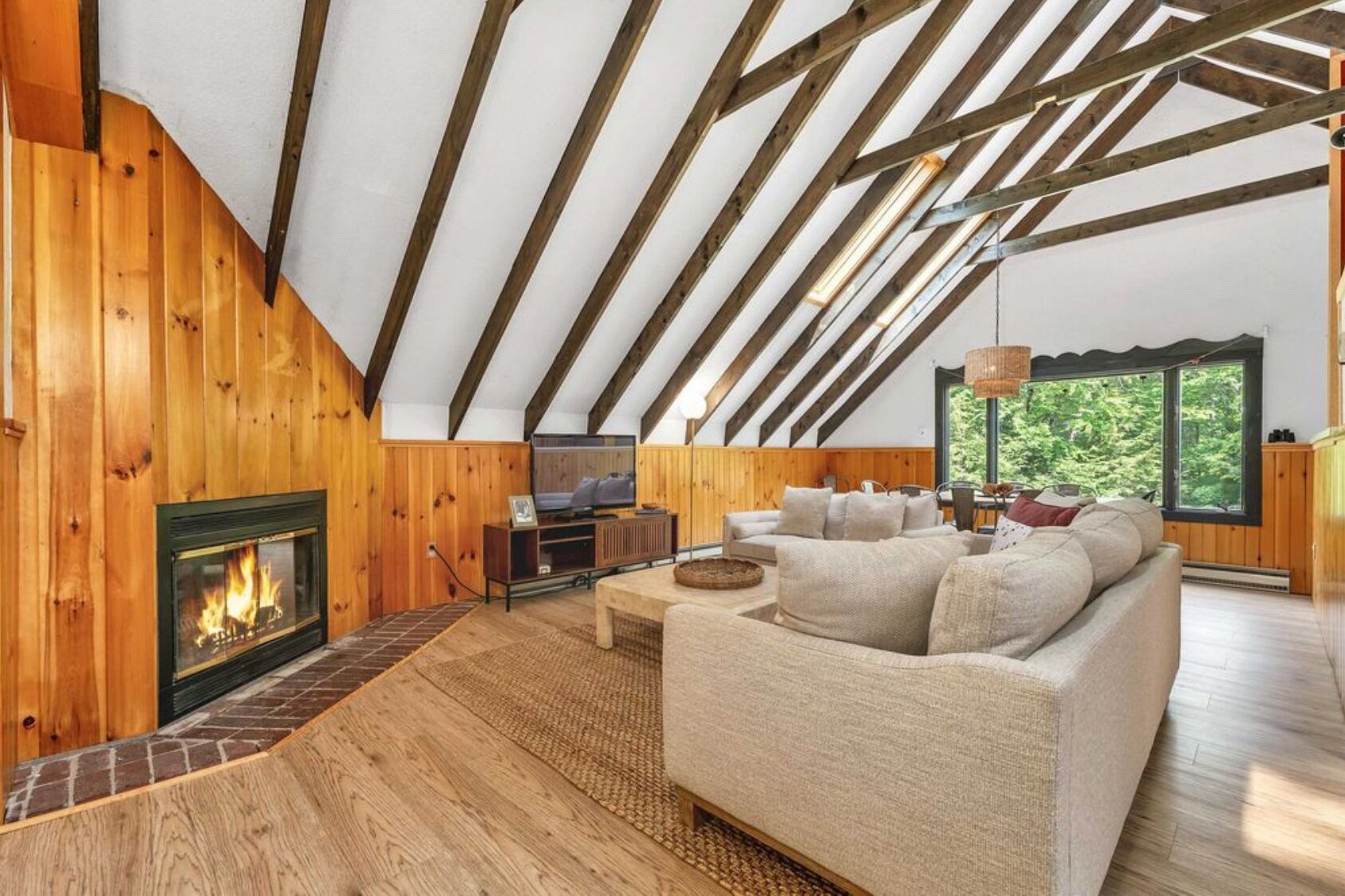 Ein gemütliches Loft mit sichtbaren Holzbalken, Kamin, einer Couch und einem großen Fenster mit Blick auf Grünflächen.
