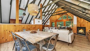 Een gezellig chalet met houten inrichting, een eethoek met tafel en stoelen, een woonkamer met open haard, en een groot raam met uitzicht op begroeiing.