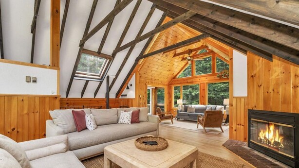 Een knus loft-woonkamer met een open haard, houten balken en grote ramen met uitzicht op de natuur.