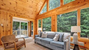 Een gezellig interieur van een blokhut met een grijze bank, een rieten stoel en een groot raam met uitzicht op het bos.