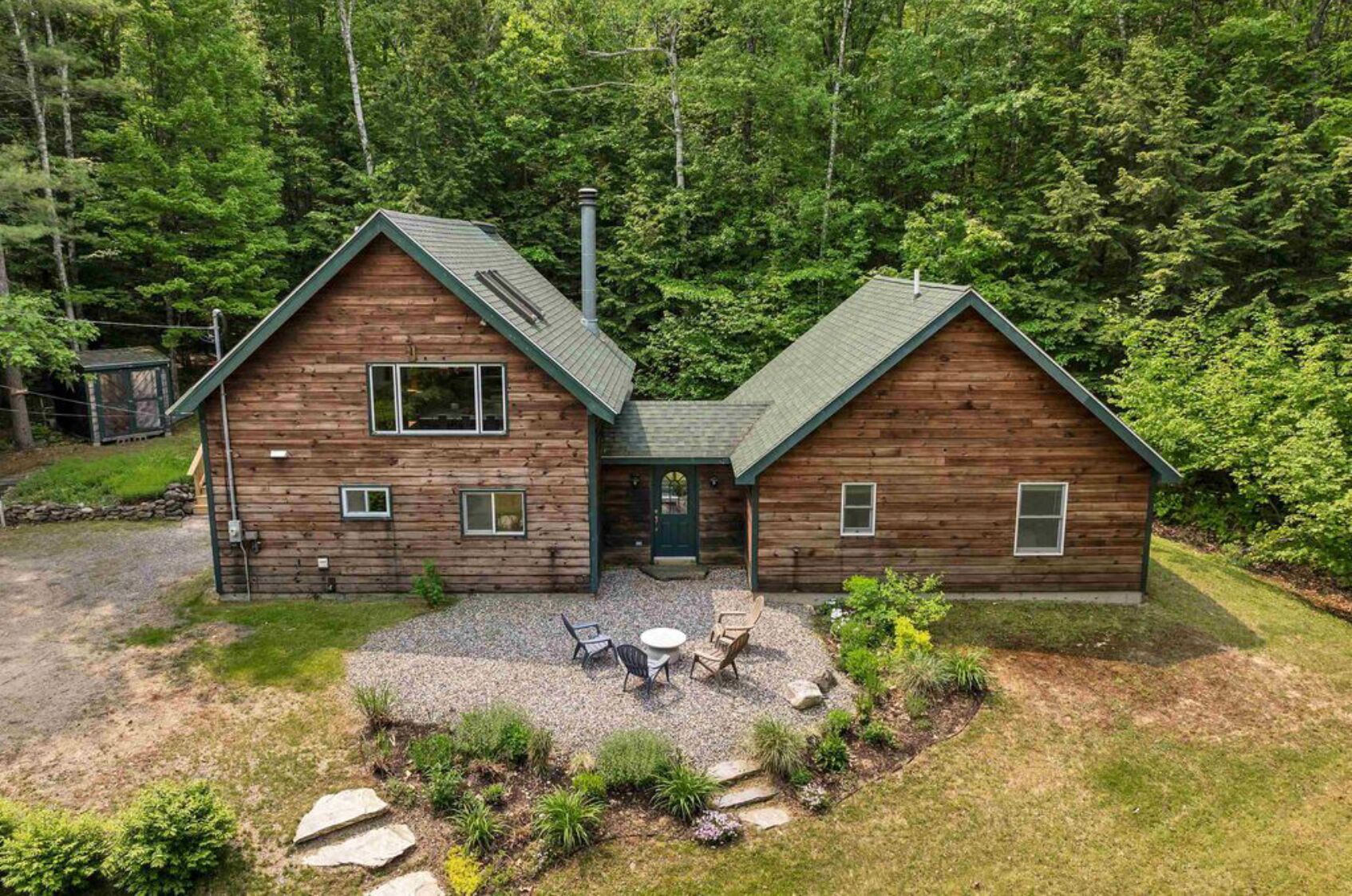 Une cabane en rondins à deux étages, avec une allée en gravier et une terrasse aménagée avec du mobilier d’extérieur.