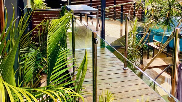 A covered outdoor seating area with a wooden deck, metal railings, and lush greenery. - The Palm Tree Loft, close to shops and lake (Green Point)