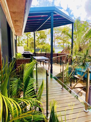 A covered outdoor seating area with a wooden deck, metal railings, and lush greenery. - The Palm Tree Loft, close to shops and lake (Green Point)