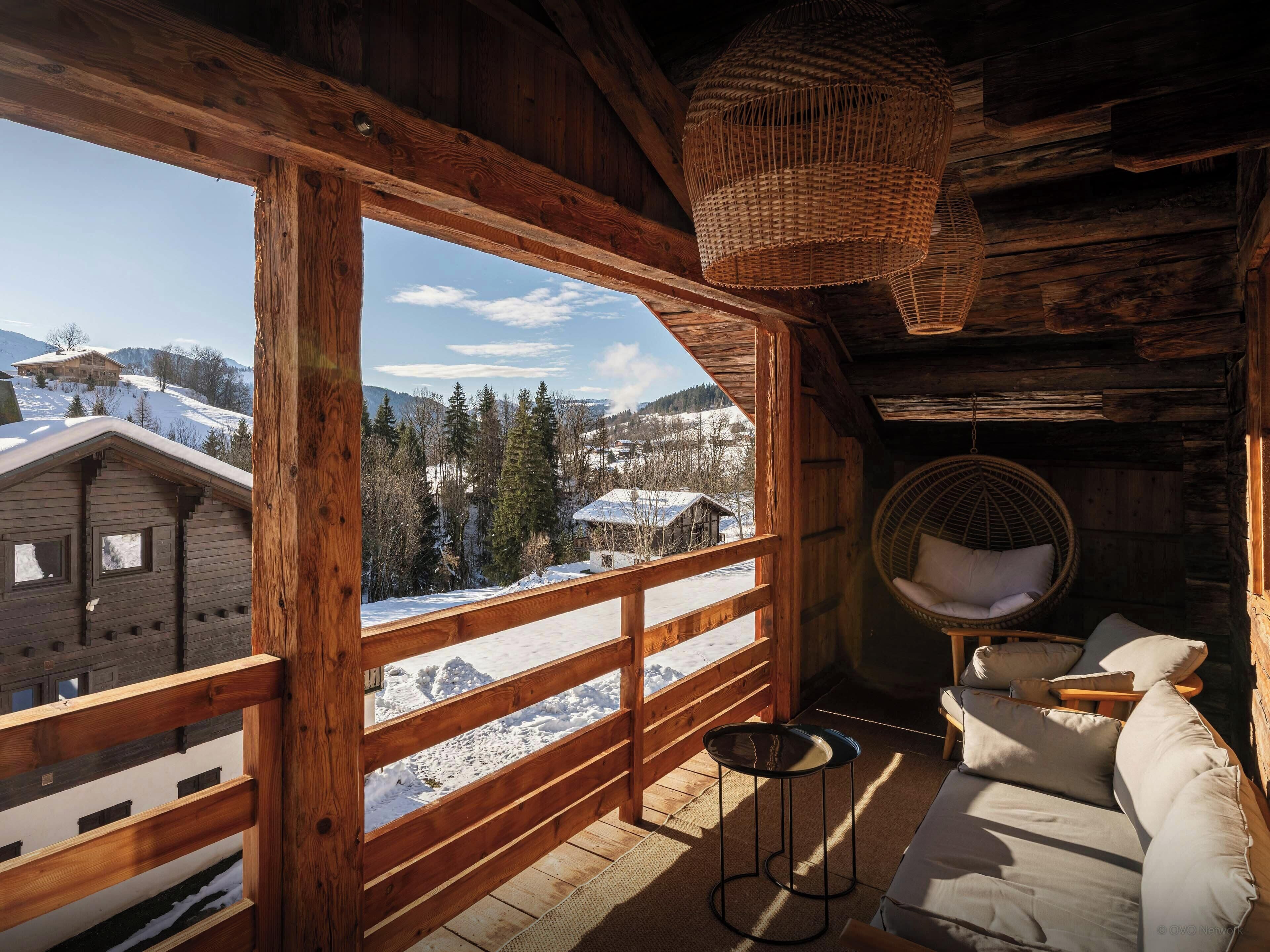 A wooden cabin with a large window offering a view of snow-covered mountains and a distant building. Inside, there is a wicker chair and a sofa with cushions.