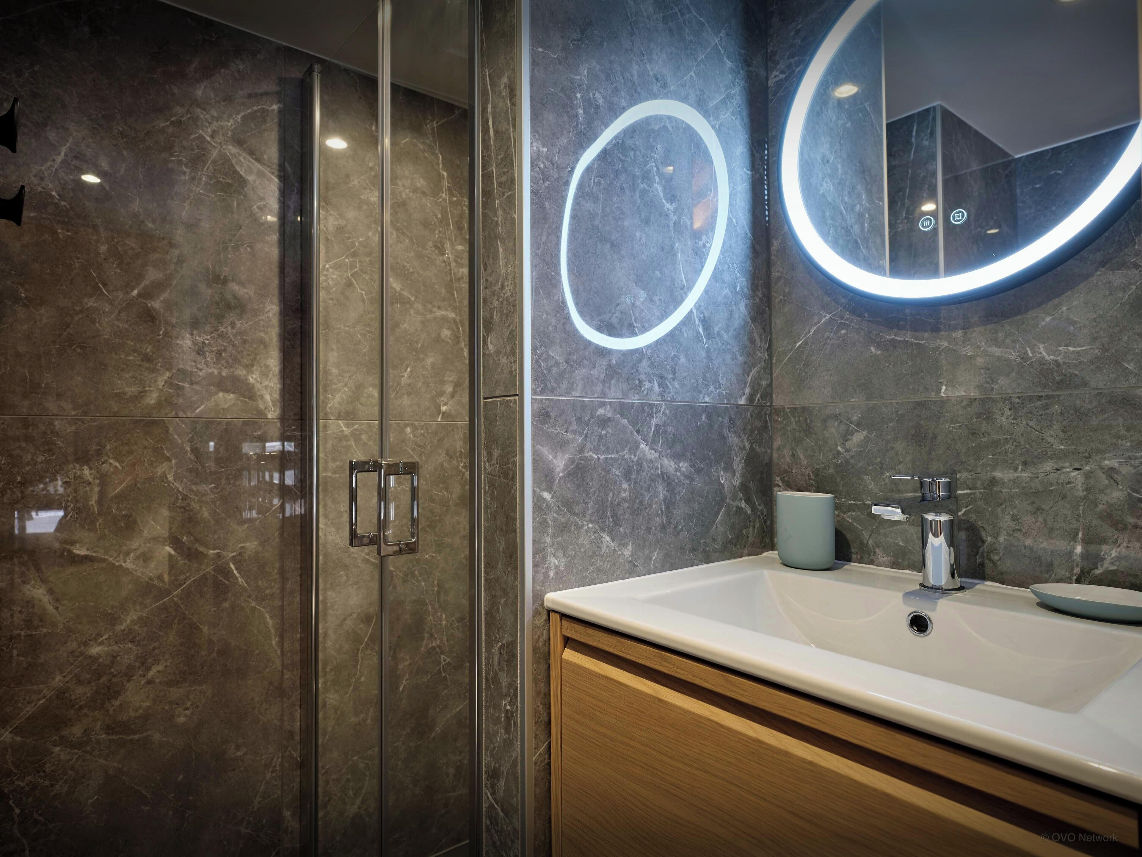 A modern bathroom with a glass shower enclosure, a marble-tiled wall, a round mirror with a light, and a white sink with a cup.