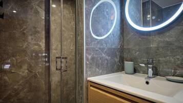 A modern bathroom with a glass shower enclosure, a marble-tiled wall, a round mirror with a light, and a white sink with a cup.