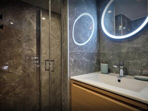 A modern bathroom with a glass shower enclosure, a marble-tiled wall, a round mirror with a light, and a white sink with a cup.