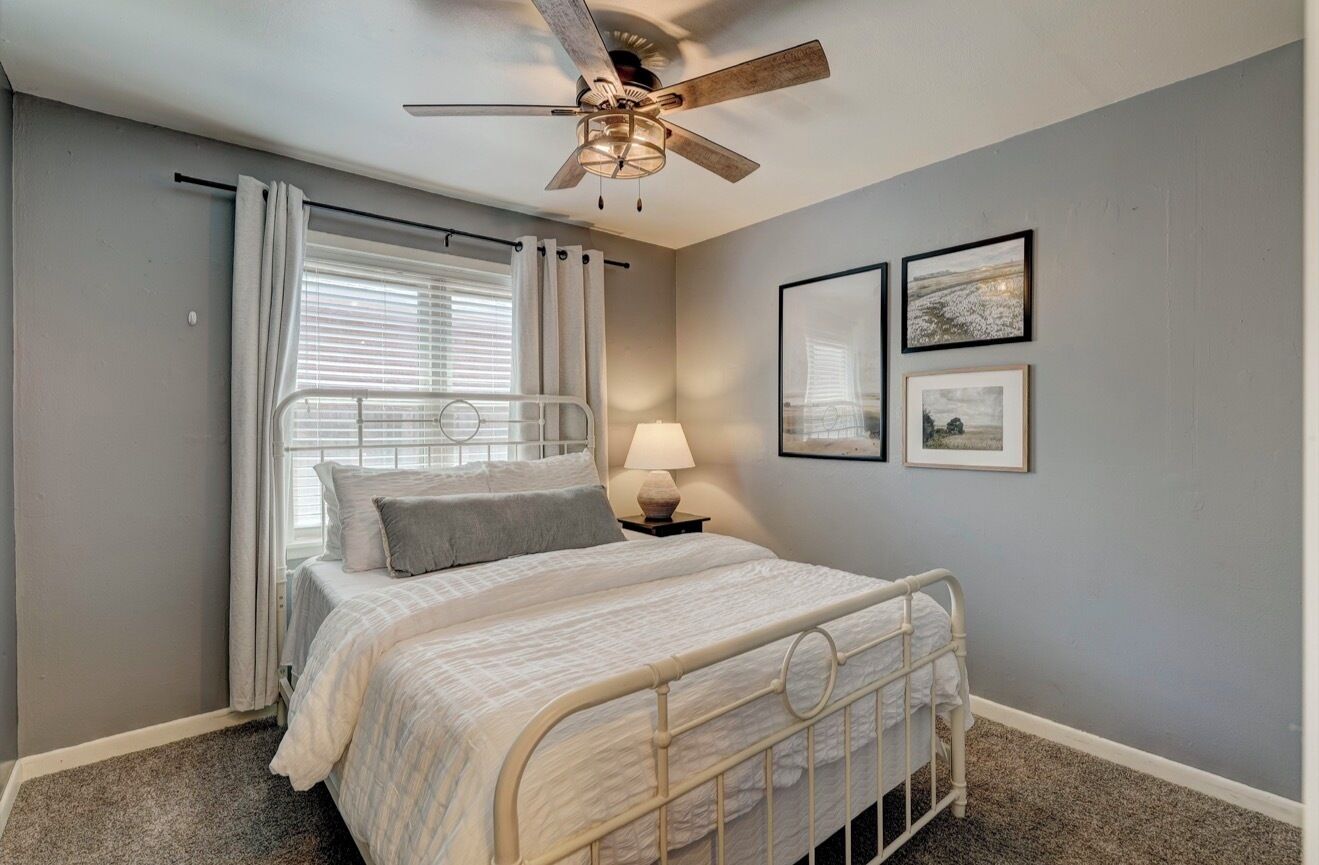 A bedroom with a bed, a ceiling fan, a window with curtains, a bedside lamp, and framed pictures on the wall.