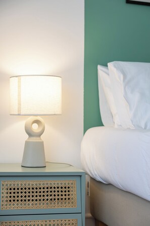 Uma cama com roupa de cama branca, uma luminária de cabeceira e uma mesinha de noite com painel decorativo.