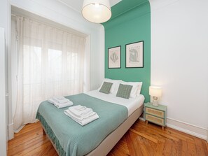 A bedroom with a bed, a nightstand, a lamp, a window with curtains, and two framed pictures on the wall.