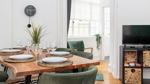 A dining area with a wooden table set for six, green chairs, a clock on the wall, and a TV on a stand.
