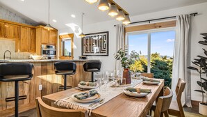 Eine moderne Küche mit Holzschränken, ein Essbereich mit Holztisch, gedeckt für vier Personen, und ein großes Fenster mit Blick ins Freie.