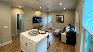 A modern kitchen with a central island, a dining area with a sofa and a TV, and a hallway leading to a bathroom.
