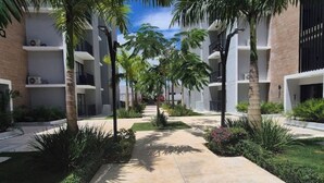 A courtyard with a central pathway flanked by palm trees and low shrubs, leading to a building with multiple balconies and a brick facade. - Caribbean home / Punta Cana /Pool, gym, playground area (Punta Cana)