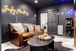 Um quarto com um sofá de couro marrom, uma mesa de centro de madeira, um letreiro neon com a inscrição 'Smoky Mountain', uma máquina de lavar e um projetor de filmes antigo.