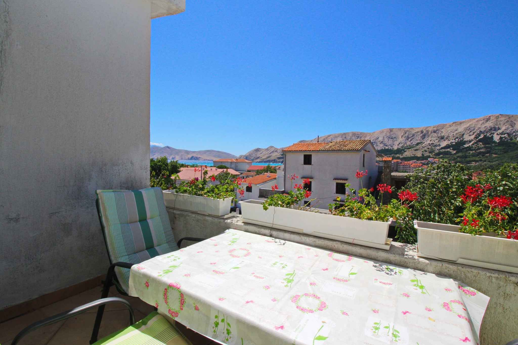 Un balcón con mesa y sillas, con vistas a un paisaje montañoso con edificios y flores rojas.