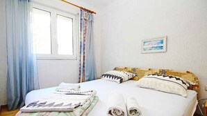 Ein ordentlich bezogenes Bett mit aufgerollten Handtüchern und Kissen, ein Fenster mit Vorhängen und ein Bild an der Wand.