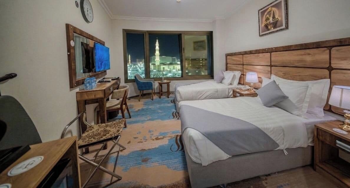 A hotel room with two beds, a desk, a chair, a TV, and a view of a cityscape at night.