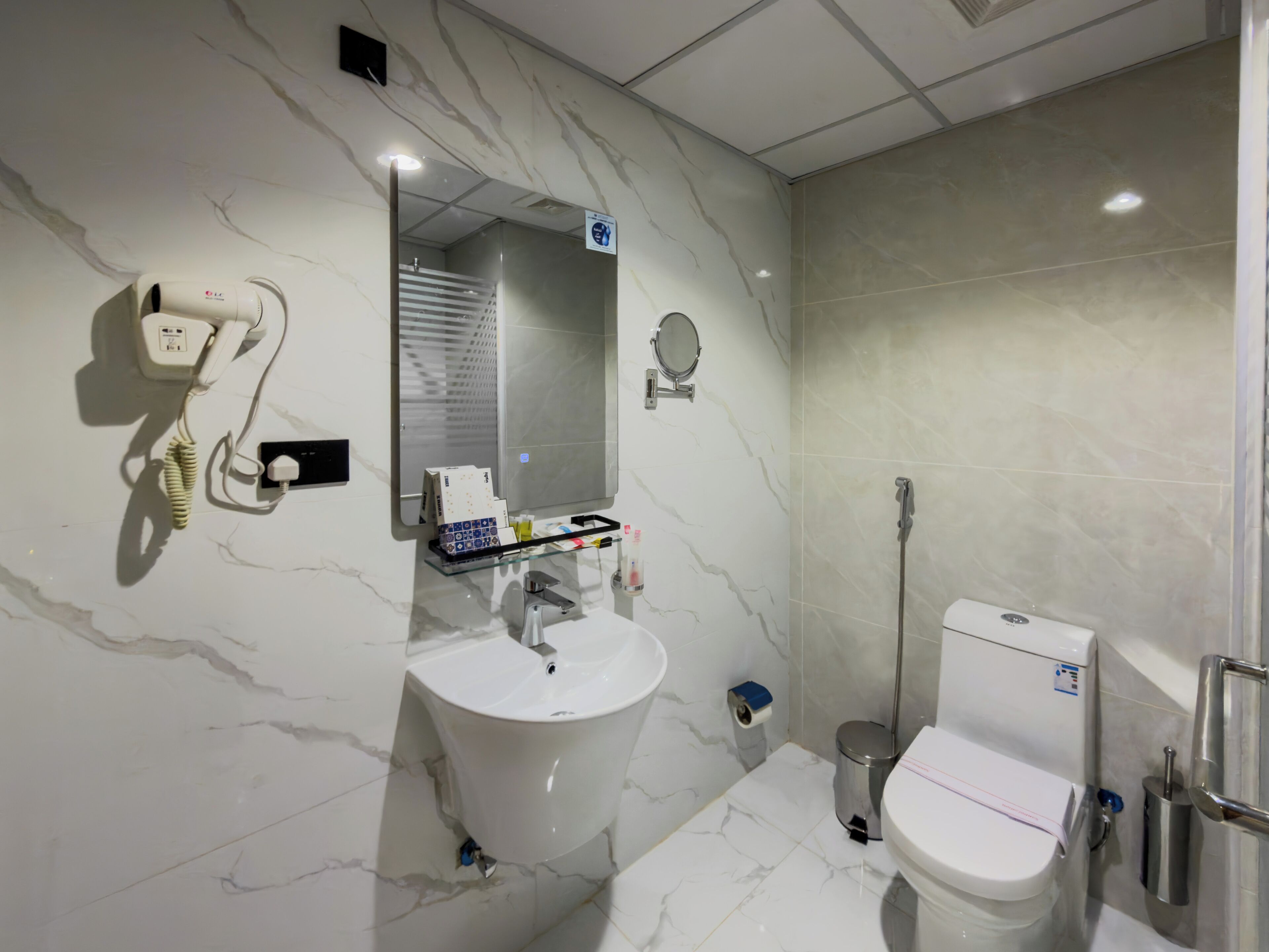 Een moderne badkamer met een marmeren wastafel, een spiegel, een haardroger, toilet en een prullenbak.