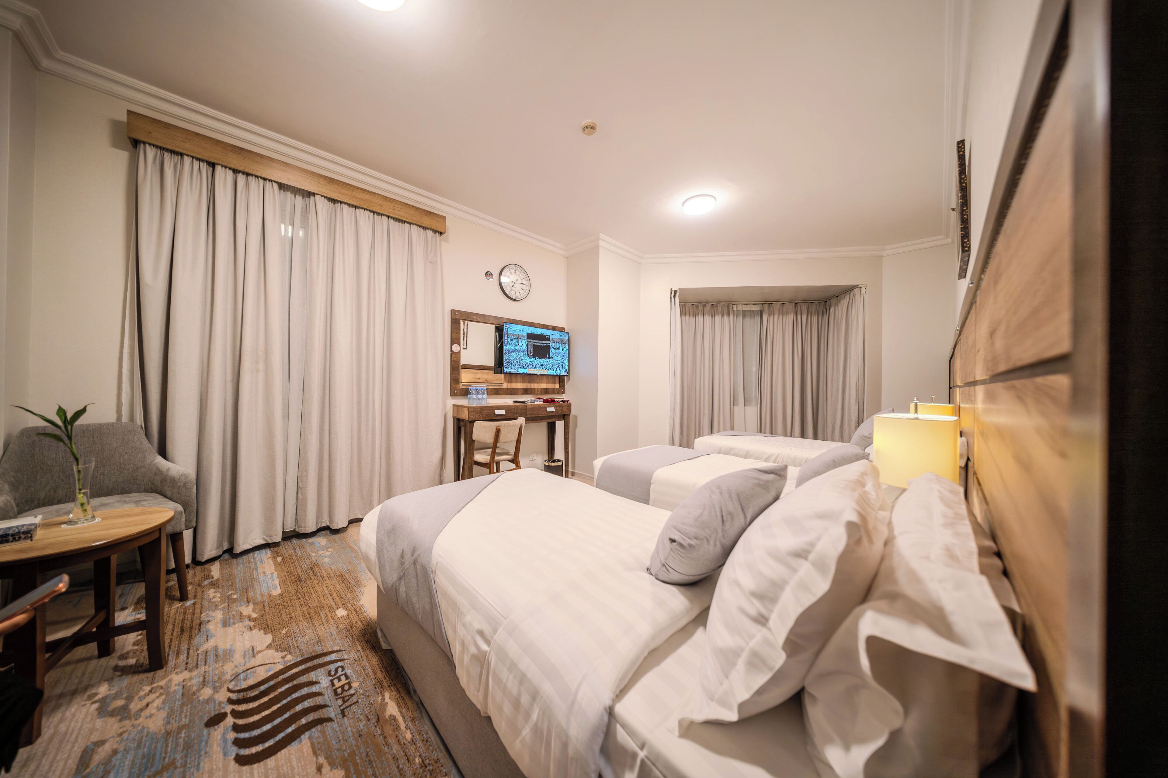 Een hotelkamer met twee bedden, een bureau met stoel, een kleintje tafeltje met een plant en een klok aan de muur.