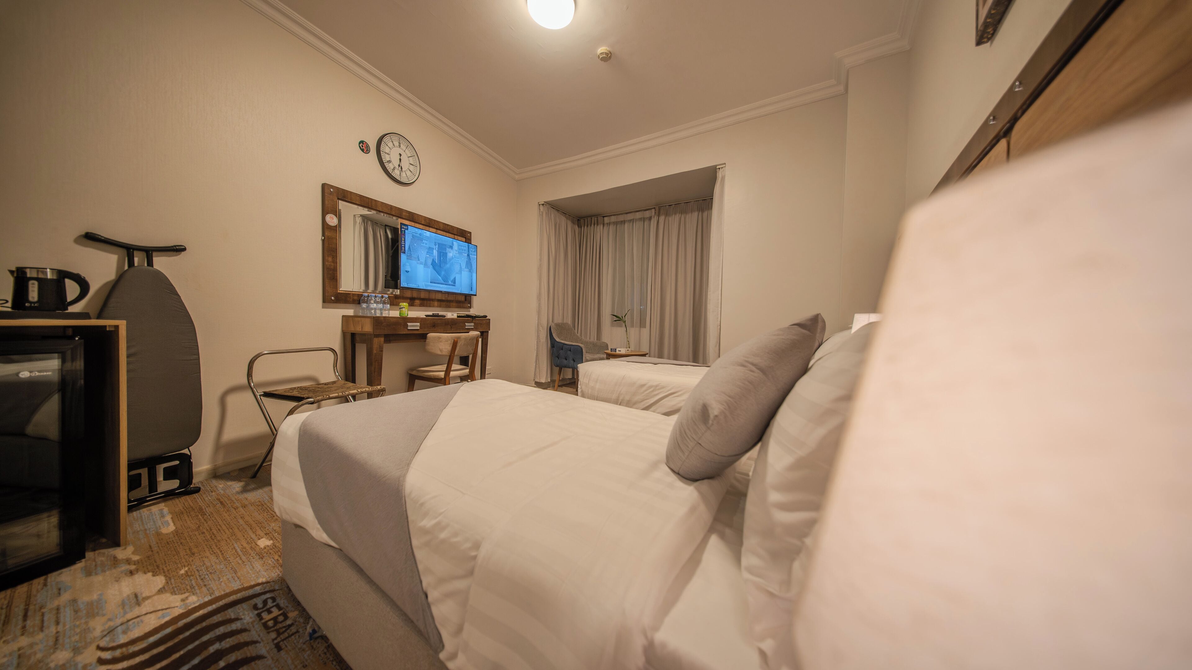 A hotel room with two beds, a desk with a computer, a chair, a clock, and a window with curtains.