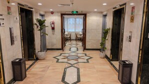 Un hall d’hôtel avec un sol en marbre, deux plantes en pot, un extincteur et un panneau indiquant la sortie.