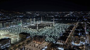 Une vaste place bien éclairée, dominée par un imposant bâtiment orné de plusieurs minarets, et entourée d’un quartier urbain densément peuplé, le tout baigné par la lumière de la nuit.