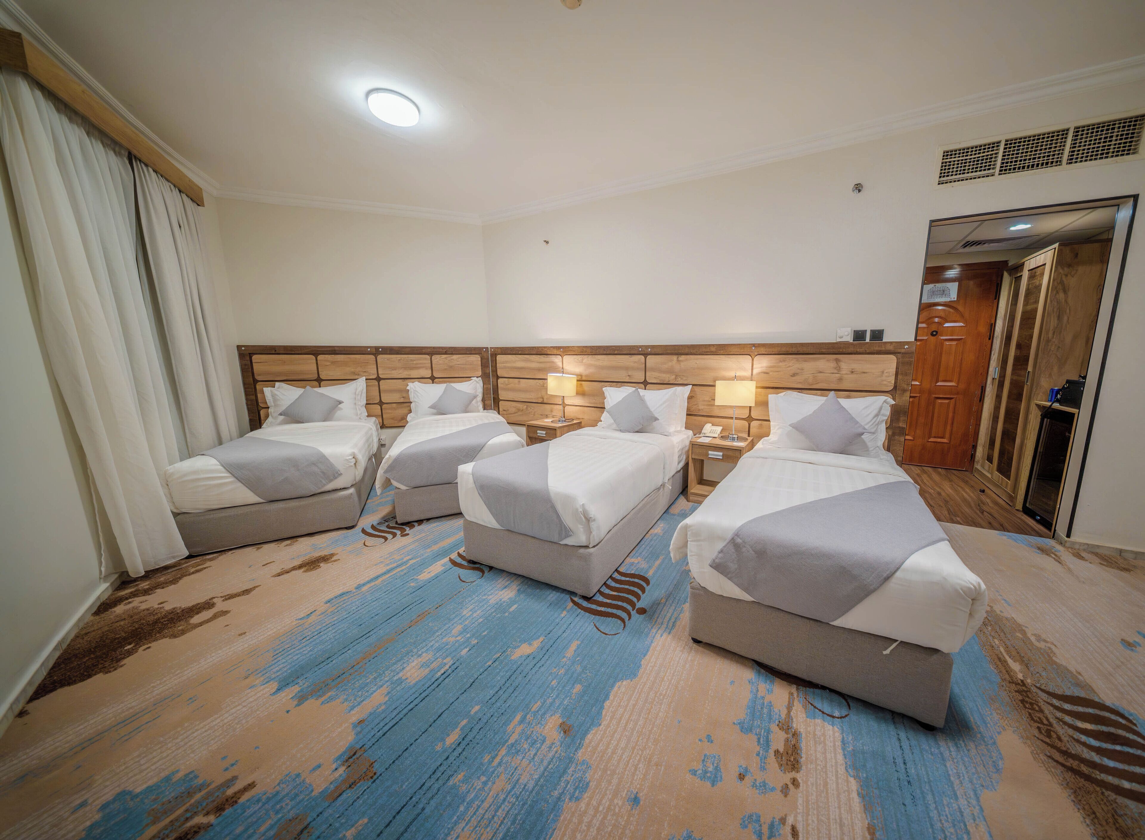 A hotel room with four beds arranged in a row, a wooden headboard, a bedside table with a lamp, and a large mirror on the wall.