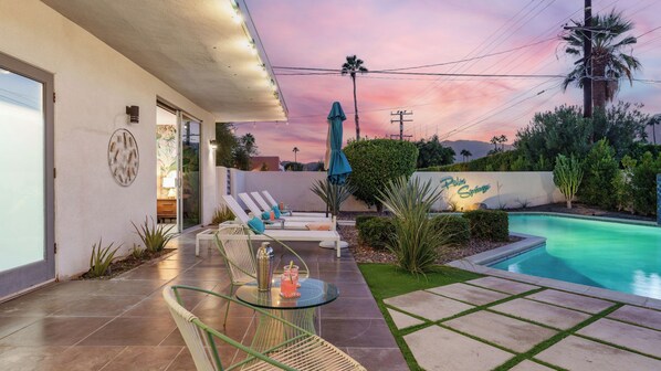 A pool area with lounge chairs and a glass table, a sign reading 'Palm Springs', and a sunset sky.