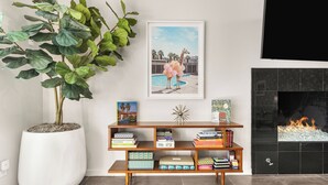 Una sala de estar con una planta grande en maceta, una estantería con libros, un cuadro enmarco y una chimenea.