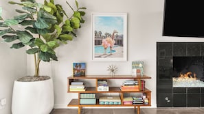 Ein Wohnzimmer mit einer großen Topfpflanze, einem Bücherregal mit Büchern, einem gerahmten Bild und einem Kamin.