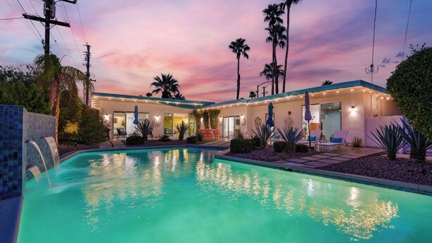 Zona junto a la piscina con piscina, palmeras y una casa con mobiliario exterior.