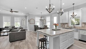 A spacious living area with a large gray sofa, a dining table, and a kitchen with white cabinets and a marble island.