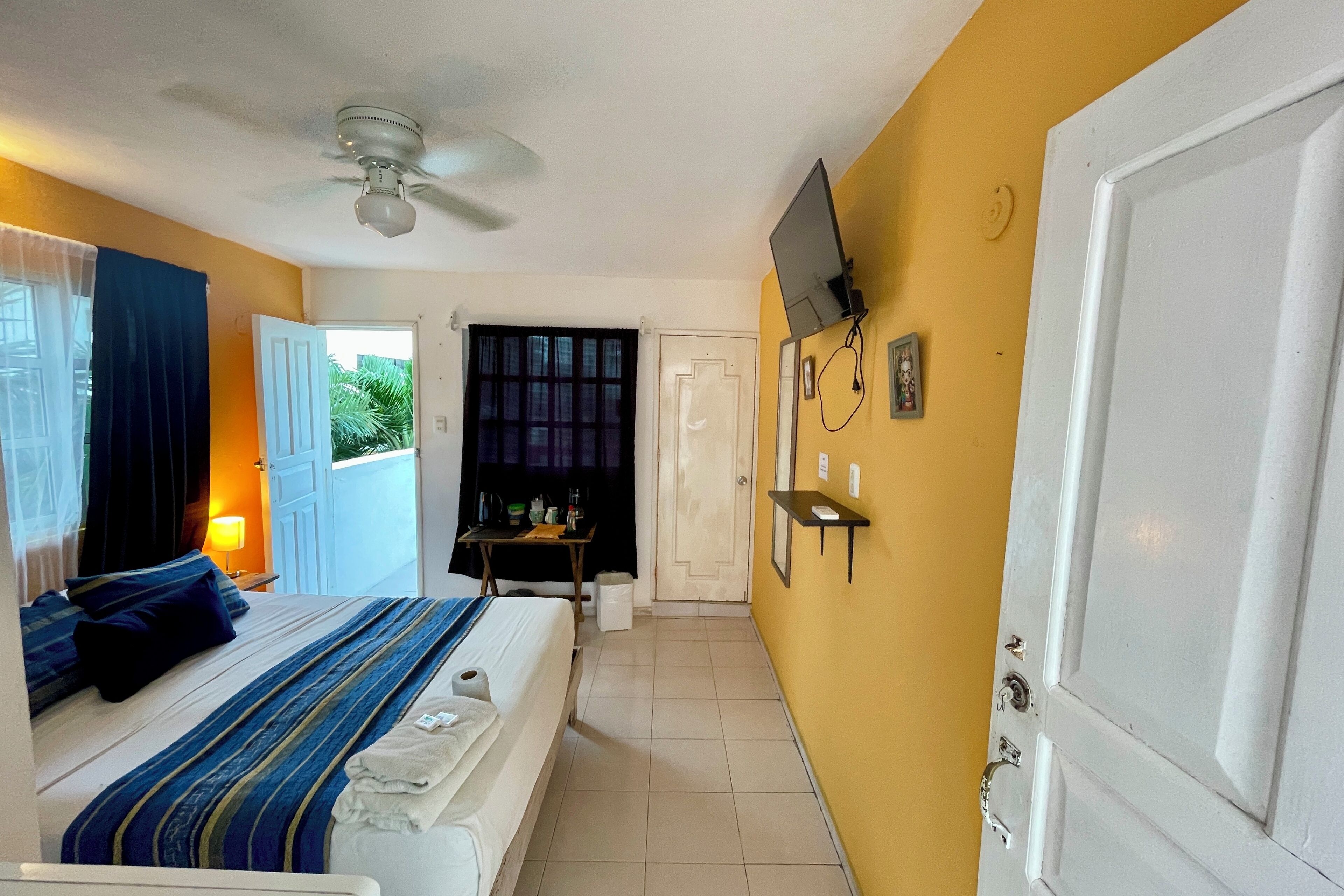 Una habitación de hotel con una cama, un ventilador de techo, un televisor montado en la pared y una puerta que da a otra habitación.
