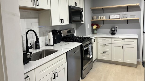 A modern kitchen with white cabinets, a stainless steel sink, a microwave, a stove, and a coffee maker. - Newly renovated 2 bedroom home located near the waterfront and Carnegie Library! (Munhall)