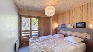 Un dormitorio con techo de madera, una cama grande con ropa blanca, una araña y vistas a árboles por una puerta corrediza.