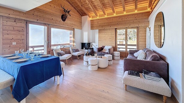 Una habitación amplia con techo alto, vigas de madera, una cabeza de ciervo montada, una mesa de comedor preparada, un sofá y una chimenea.