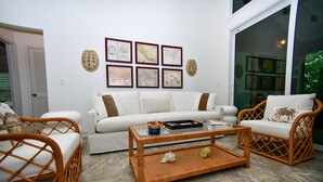 Uma sala de estar com um sofá branco, duas cadeiras de vime, uma mesa de centro de vidro e quadros na parede.