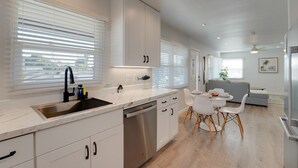 Eine moderne Küche mit weißen Schränken, einer Edelstahl-Spüle und einem Essbereich mit grauem Sofa und weißen Stühlen.