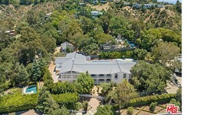 An aerial view of a large, modern house with a swimming pool, surrounded by dense greenery and other houses on a hillside.