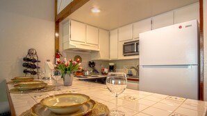 Una cocina con armarios blancos, un microondas y un refrigerador. Hay platos, una copa de vino y un florero con flores en la encimera.