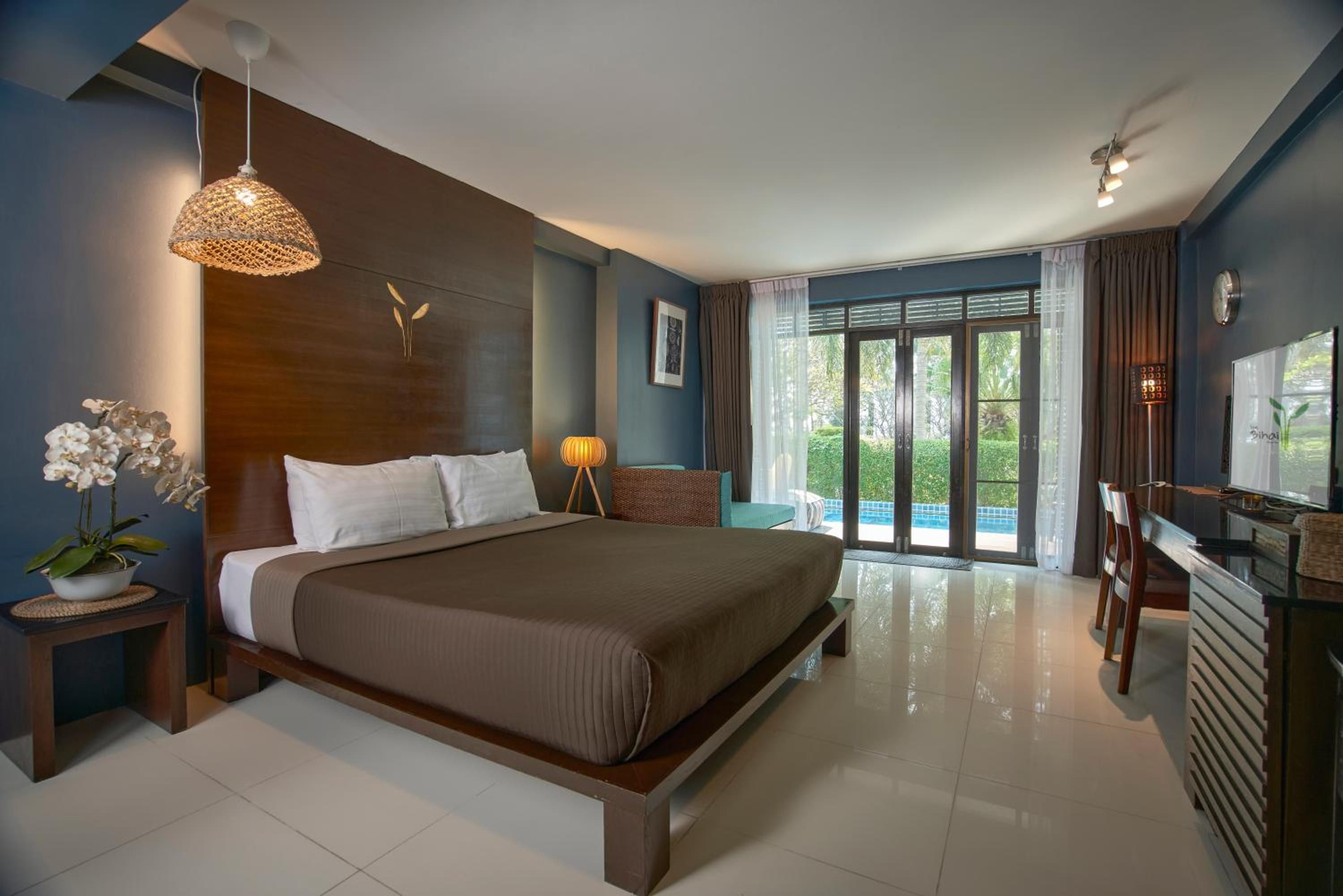 A modern hotel room with a large bed, a desk, a chair, a potted plant, and a view of the outdoors.