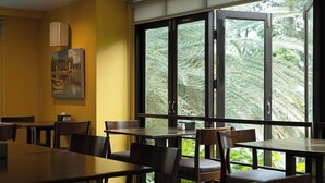 Un restaurant avec des tables et des chaises en bois, de grandes fenêtres donnant sur des palmiers et un tableau accroché au mur.